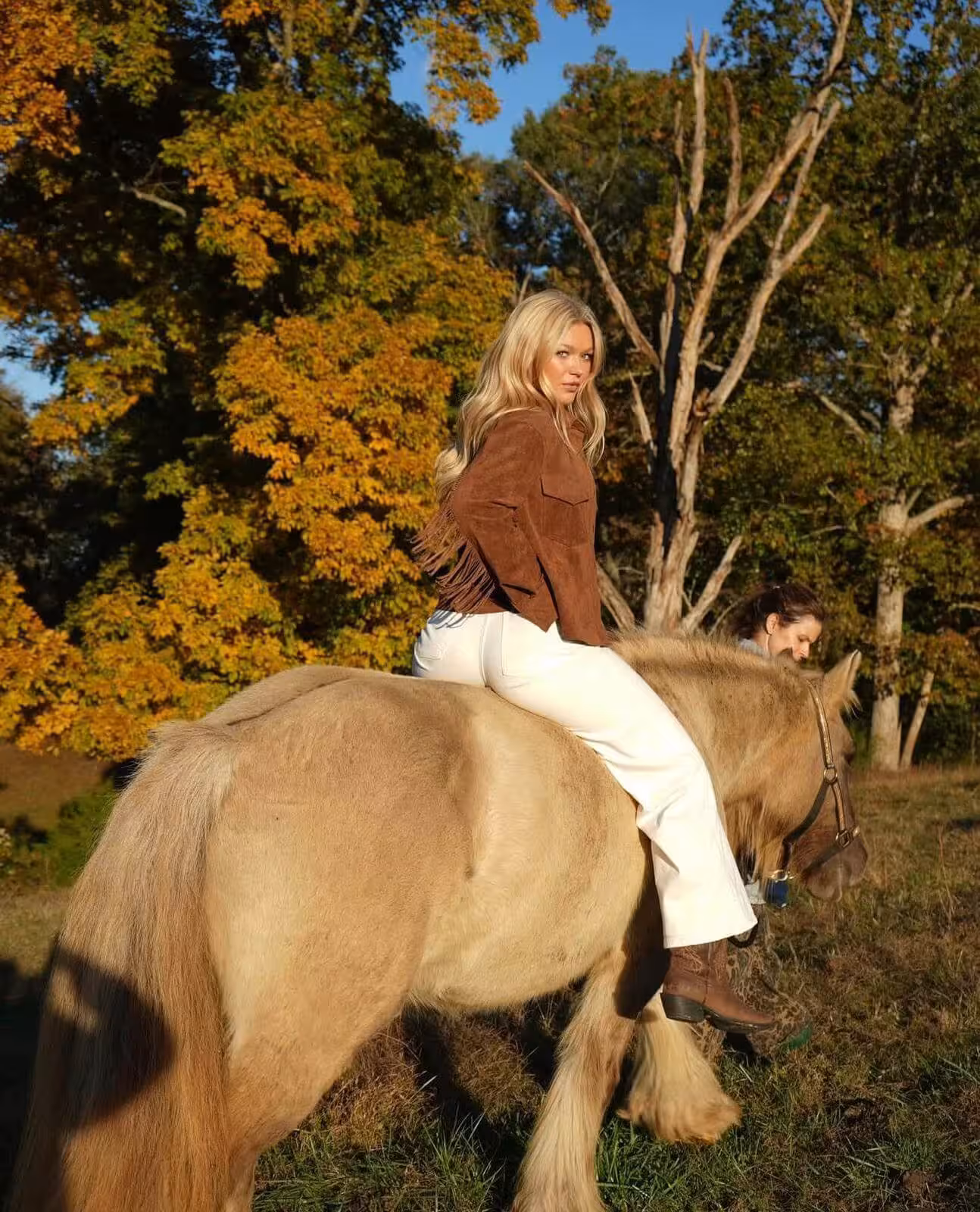 Katie Sigmond in brown jacket and white pants sitting sideways on a light brown horse in a sunlit autumn landscape.