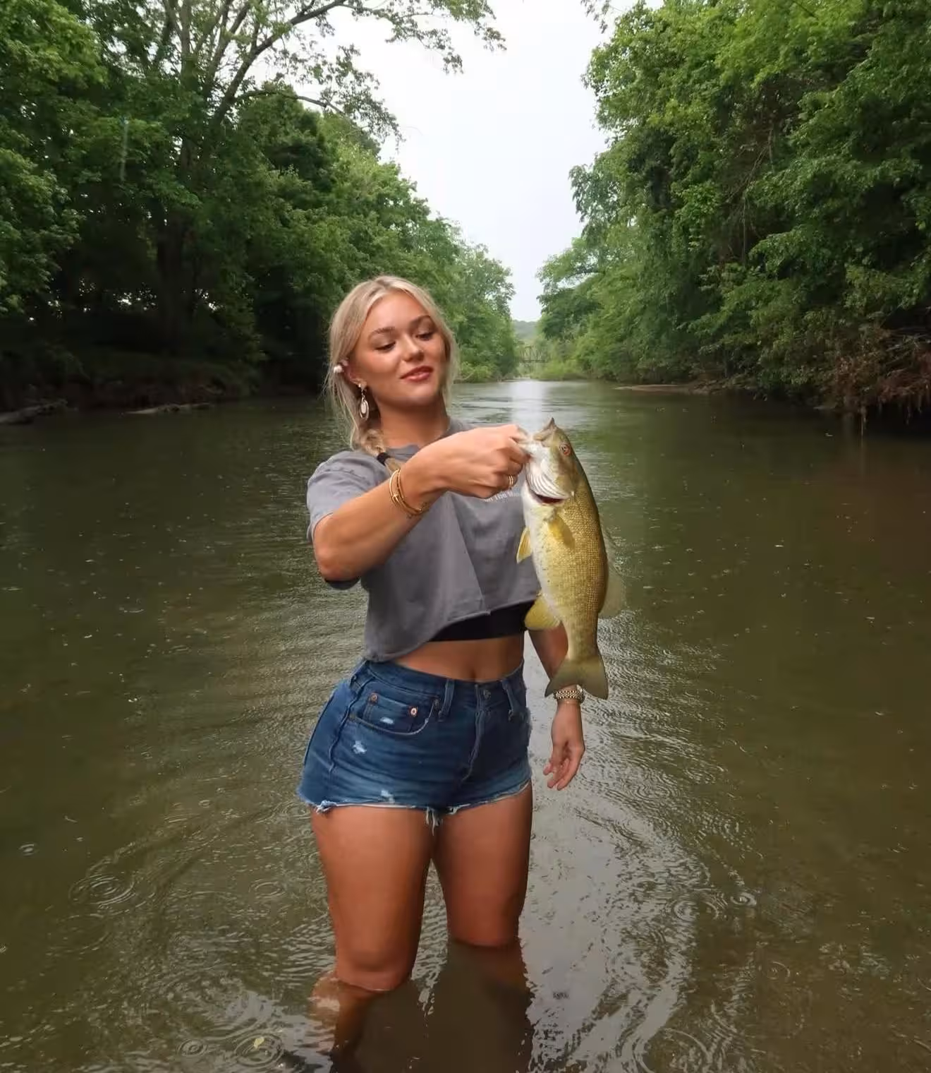 Katie Sigmond standing knee-deep in a river holding a large fish with green trees lining the riverbanks.